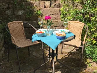 Ein gemütlicher Tisch im Freien mit zwei Stühlen. Auf dem Tisch stehen Gläser, eine Blumenvase und Schalen mit Snacks.