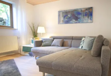 Ein gemütliches Wohnzimmer mit einer grauen Couch und einem farbigen Bild an der Wand. Eine Lampe und eine Pflanze verleihen dem Raum eine einladende Atmosphäre.