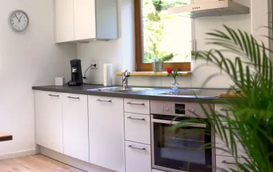 Eine moderne Küche mit weißen Schränken und einem großen Fenster. Auf der Arbeitsplatte stehen eine Kaffeemaschine und ein kleiner Blumenstrauß.