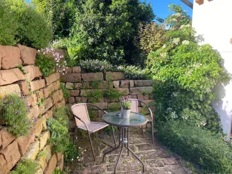 Ein kleiner, grüner Garten mit einer gemütlichen Sitzecke. Der Tisch ist aus Glas, und es gibt zwei Stühle, umgeben von Pflanzen und Blumen.