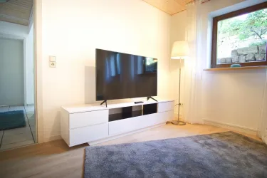 Ein modernes Wohnzimmer mit einem Flachbildfernseher auf einem weißen TV-unterschrank. Das Zimmer ist hell und freundlich, mit einem weichen Teppich und einem Fenster, das Tageslicht hineinlässt.