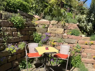 Ein gemütlicher Gartenbereich mit zwei Stühlen und einem kleinen Tisch. Umgeben von bunten Blumen und grünen Pflanzen.