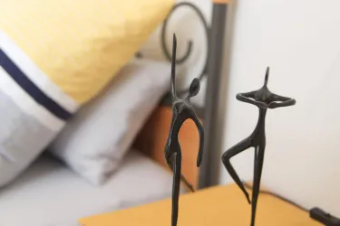 Zwei schwarze Skulpturen von tanzenden Figuren stehen auf einem Tisch. Im Hintergrund sind Bettwäsche und Kissen sichtbar.