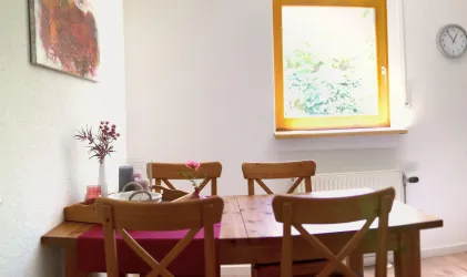 Ein gemütlicher Essbereich mit einem Holztisch und vier Stühlen. Ein Fenster mit gelben Rahmen lässt viel natürliches Licht herein.