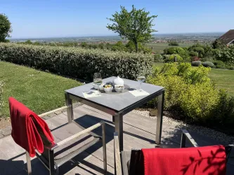 Ein Tisch mit Gläsern und einer Schüssel auf einer Terrasse mit Blick auf eine grüne Landschaft. Zwei Stühle mit roten Decken stehen bereit, um die Aussicht zu genießen.