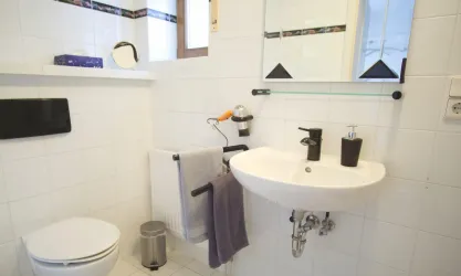 Ein modernes Badezimmer mit weißen Fliesen und einem Waschbecken. Es gibt einen Spiegel, eine Toilette und Handtücher, die ordentlich aufgehängt sind.