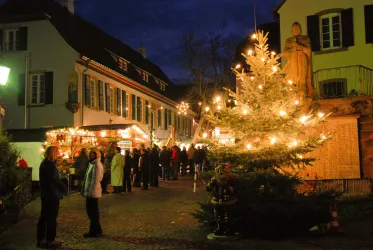 Ein festlicher Weihnachtsmarkt mit einem beleuchteten Tannenbaum. Menschen stehen zusammen und genießen die festliche Stimmung in einer malerischen Gasse.