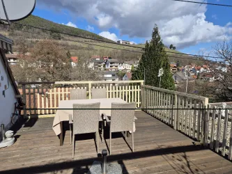 Eine Holzterrasse mit einem Tisch und Stühlen steht in einer ländlichen Umgebung. Im Hintergrund sind Häuser und eine bewaldete Hügelkette unter einem teilweise bewölkten Himmel zu sehen.