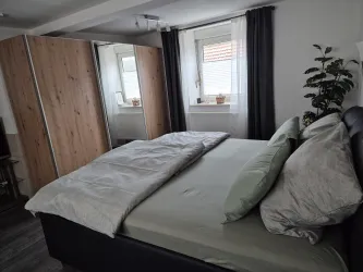 Ein modernes Schlafzimmer mit einem großen Bett und zwei Kissen. Im Hintergrund gibt es einen Kleiderschrank und eine Pflanze vor einem Fenster.