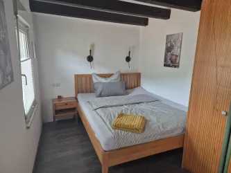 Ein gemütliches Schlafzimmer mit einem Holzbett und weichen Kissen. An der Wand hängen zwei Lampen und ein Bild, während ein Fenster Tageslicht hereinscheinen lässt.