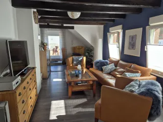 Ein gemütliches Wohnzimmer mit einer braunen Couch, einem Holztisch und einem Fernseher. Helle Wände und Fenster sorgen für eine einladende Atmosphäre.