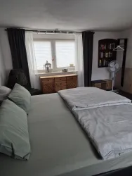 Ein gemütliches Schlafzimmer mit einem großen Bett und grünen Kissen. An der Wand steht ein Bücherregal und ein Fenster mit Vorhängen.