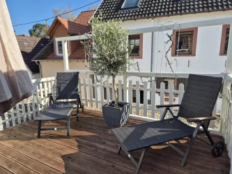 Eine Terrasse mit zwei Liegestühlen aus Holz und einem Blumentopf. Im Hintergrund sind Häuser und ein blauer Himmel sichtbar.