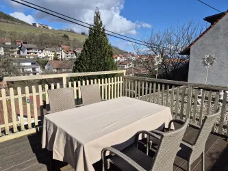 Ein gemütlicher Balkon mit einem Tisch und Stühlen. Im Hintergrund sind Häuser und eine grüne Landschaft sichtbar.