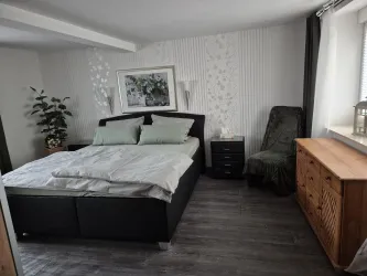 Ein modernes Schlafzimmer mit einem großen Bett und sanften grünen Bettdecken. An der Wand hängt ein dekoratives Bild und es gibt eine gemütliche Sitzecke.