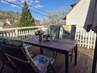 Ein einladender Balkon mit einem Holztisch und zwei Stühlen. Auf dem Tisch stehen Blumen und ein Flasche Wein, umgeben von einer schönen Landschaft.