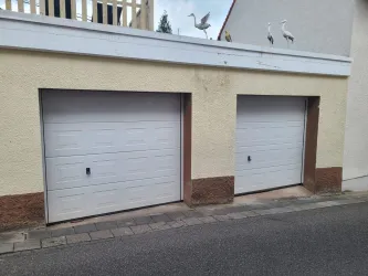 Zwei weiße Garagentore an einer Wand. Über den Toren stehen einige Störche.