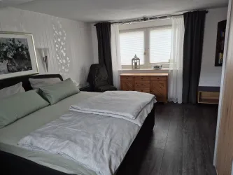 Ein modern eingerichtetes Schlafzimmer mit einem großen Bett und frischen Bettwaren. Es gibt ein Fenster mit Vorhängen und einen Holztisch mit einer dekorativen Laterne.