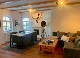 Ein gemütliches Wohnzimmer mit einem großen Esstisch und Stühlen. Der Raum hat große Fenster, ein Sofa und eine dekorative Wand mit einem Geweih.