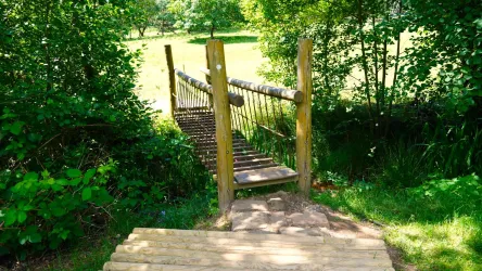 Eine kleine Holzbrücke führt über ein schmalen Abgrund. Umgeben von grünen Bäumen und Wiese wirkt die Szenerie naturverbunden und einladend.