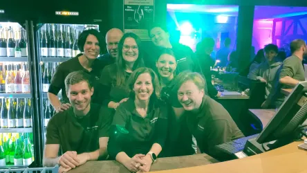 Eine Gruppe von fröhlichen Menschen steht hinter einer Bar. Sie tragen alle ähnliche braune T-Shirts und lächeln in die Kamera.