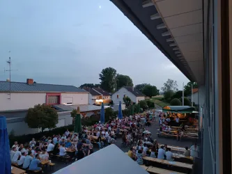 Ein belebter Außenbereich mit vielen Tischen und Gästen, die zusammen essen und trinken. Die Atmosphäre ist entspannt und gesellig, umgeben von Gebäuden und Bäumen.