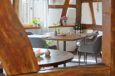 Ein gemütlicher Essbereich mit einem Holztisch und modernen Stühlen. Im Hintergrund sind Pflanzen und Flaschen sichtbar.