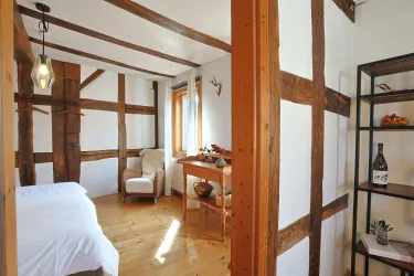 Ein gemütliches Zimmer mit Holzbalken und hellen Wänden. An einem Tisch stehen Dekorationsgegenstände, und ein Sessel lädt zum Entspannen ein.