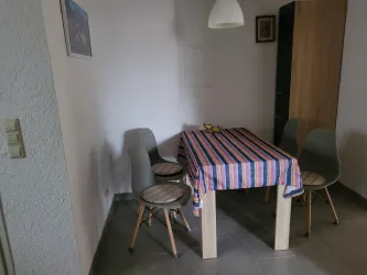 Ein einfacher Essbereich mit einem Holztisch und buntem Tischdecken. Um den Tisch stehen vier Stühle, und es gibt einen Schrank im Hintergrund.
