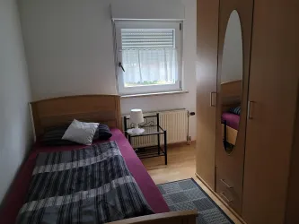 Ein kleines Schlafzimmer mit zwei Einzelbett und einem Nachttisch. Der Raum hat einen Schrank und ein Fenster mit Vorhängen.