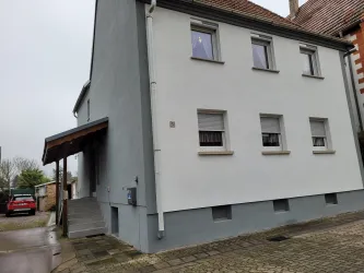 Ein modernes, graues Wohnhaus mit einem überdachten Eingangsbereich. Der Himmel ist bewölkt und die Umgebung zeigt gepflasterte Wege.