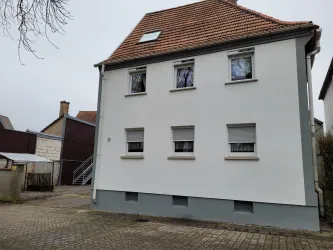 Ein modernes Wohnhaus mit einer hellen Fassade und einem roten Satteldach. Im Vordergrund ist ein gepflasterter Parkplatz zu sehen.