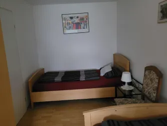 Ein schlichtes Schlafzimmer mit zwei Einzelbetten, einem Nachttisch und einem Stuhl. An der Wand hängt ein gemaltes Bild.