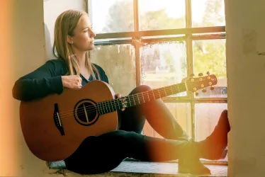 Eine Frau sitzt am Fenster und spielt Gitarre. Die natürliche Beleuchtung schafft eine warme und inspirierende Atmosphäre.