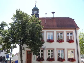 Ein hübsches Gebäude mit einer Uhr und bunten Blumenkästen. Vor dem Haus steht ein großer Baum.