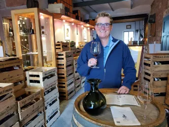 Eine Person steht in einer Weinlounge und hält ein Weinglas in der Hand. Im Hintergrund sind Holzregale mit Weinflaschen und Kisten zu sehen.