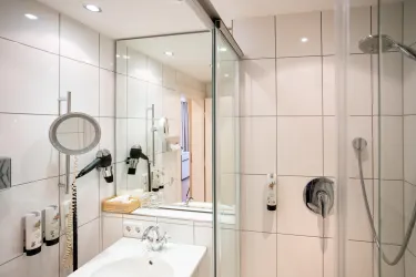 Ein modernes Badezimmer mit einer Dusche und einem Waschbecken. Die Wände sind mit weißen Fliesen gefliest und es gibt ein großes Spiegel.
