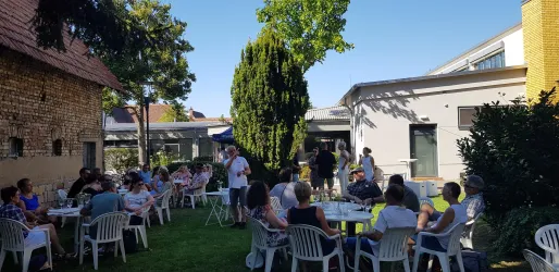 Eine fröhliche Gartenparty mit vielen Gästen, die an Tischen sitzen. Die Sonne scheint, und im Hintergrund sind Bäume und Gebäude sichtbar.