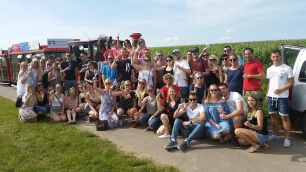 Eine große Gruppe von Menschen posiert fröhlich im Freien. Im Hintergrund sind Weinreben und ein roter Wagen zu sehen.