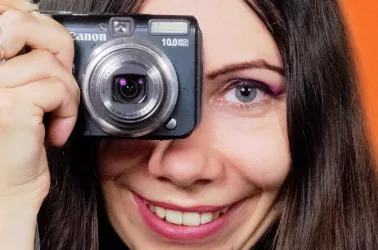 Eine Frau hält eine Kamera vor ihr Gesicht und lächelt. Sie hat lange, dunkle Haare und trägt einen farbigen Lidschatten.