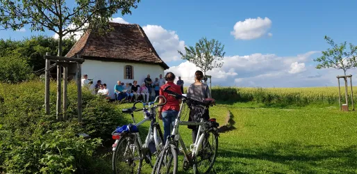 Zwei Personen stehen mit Fahrrädern auf einer Wiese vor einer Kapelle. Im Hintergrund sitzen mehrere Menschen und genießen die Landschaft.