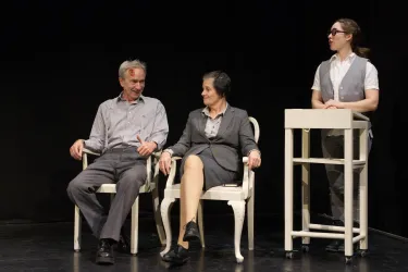 Zwei Personen sitzen auf Stühlen, während eine dritte Person am Tisch steht. Es scheint eine Szene aus einem Theaterstück zu sein.