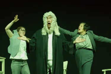 Eine Bühnenaufführung mit drei Schauspielern. Der Mann in der Mitte trägt eine Richterrobe und eine Perücke, während die beiden Frauen dramatisch agieren.