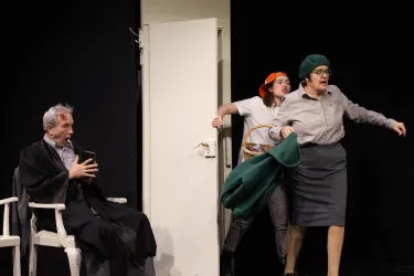 Eine Theaterszene mit drei Darstellern. Ein Mann sitzt auf einem Stuhl, während zwei Frauen lebhaft miteinander interagieren.