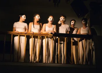 Sechs Personen stehen auf einem Balkon und tragen weiße Kleidung sowie Masken. Die Atmosphäre ist geheimnisvoll und dramatisch.