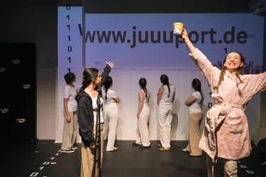 Eine Gruppe von Personen steht auf der Bühne, während eine Person fröhlich eine Tasse hochhält. Im Hintergrund ist eine Webseite mit der Adresse "www.juuuport.de" sichtbar.
