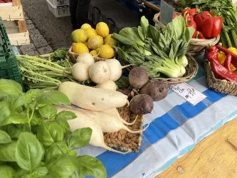Ein frischer Marktstand mit einer Auswahl an Gemüse und Obst. Darunter sind Radieschen, Paprika, Zwiebeln und Zitrone zu sehen.