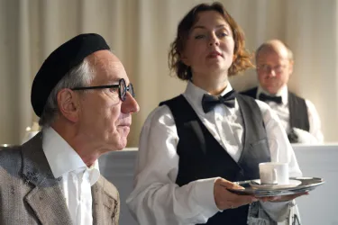 Eine Szene aus einem Restaurant mit einem älteren Mann und einer Bedienung, die ein Tablett mit einer Tasse serviert. Im Hintergrund sitzt ein weiterer Mann.