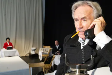 Ein Mann spricht am Telefon und trägt einen Stift im Mund. Im Hintergrund sitzen zwei Personen an Tischen.