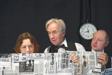 Drei Personen betrachten intensiv ein großes Stück Zeitungspapier. Im Hintergrund steht eine Uhr, die die Zeit anzeigt.